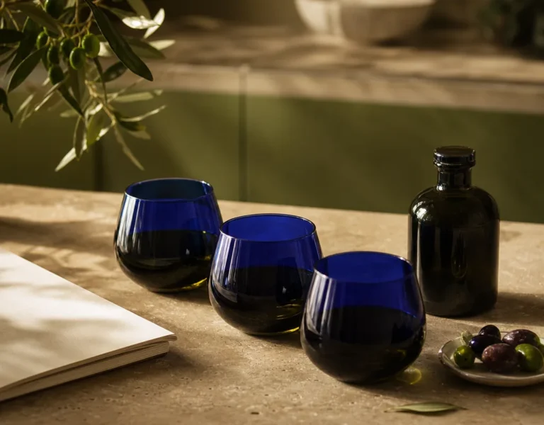 Blue olive oil tasting glasses on a light table beside olives and an unlabelled bottle